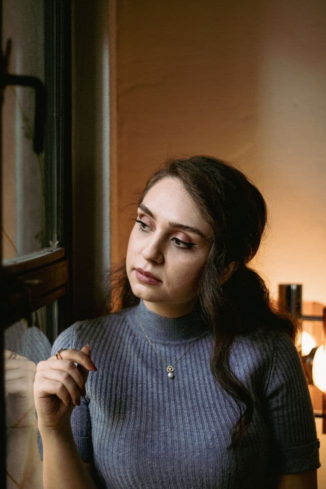 A young woman in a blue top looking thoughtfully out of a window