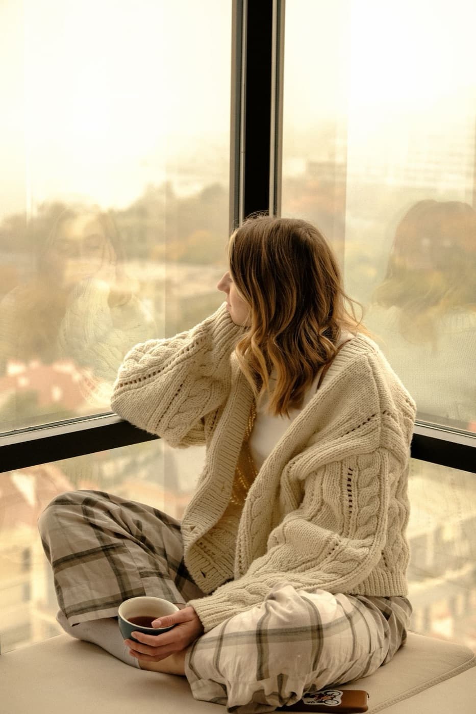 A woman in a cream jumper looking out of a window with a hot drink