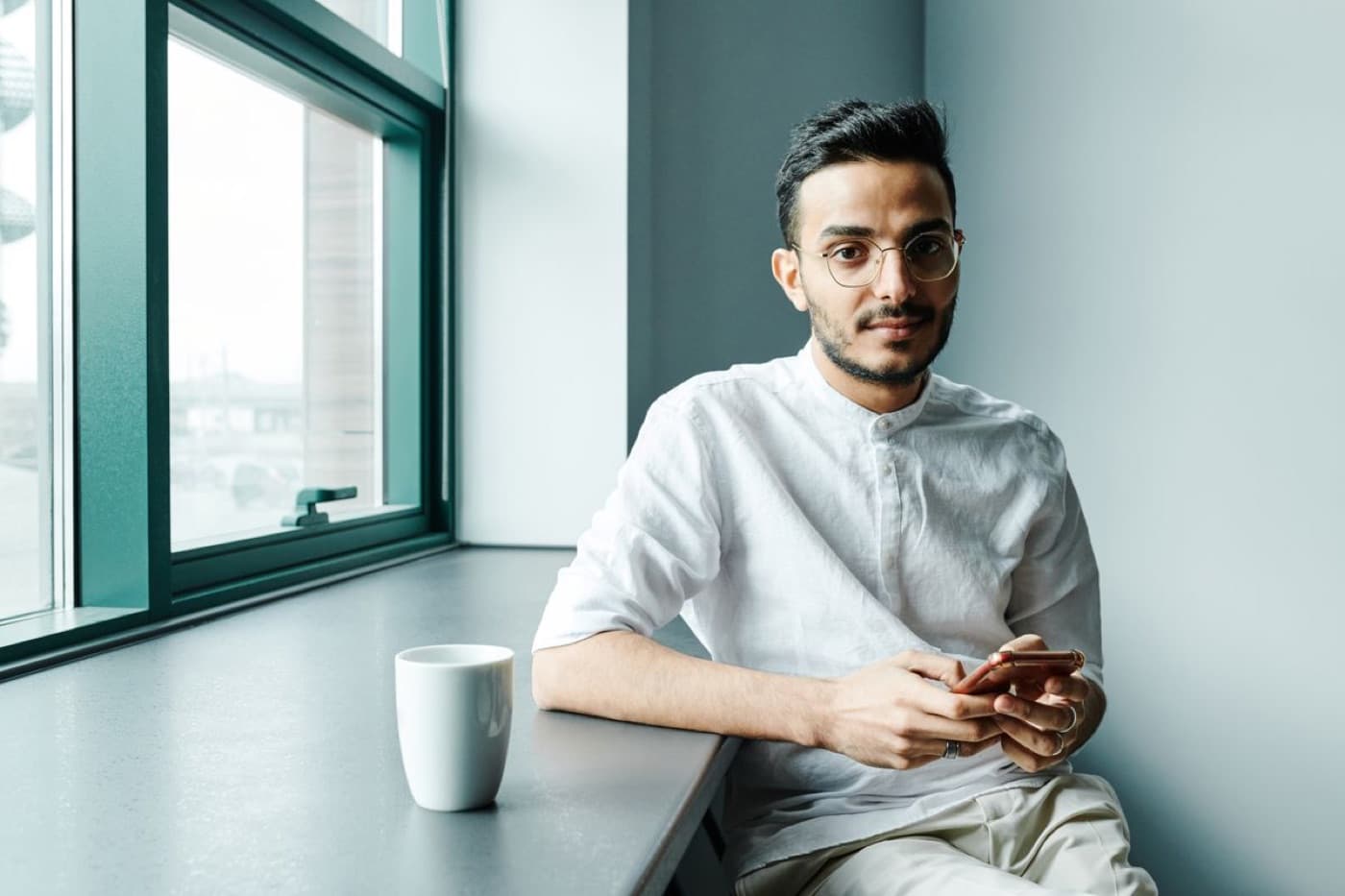 A man sitting by a bright window with a coffee and his phone