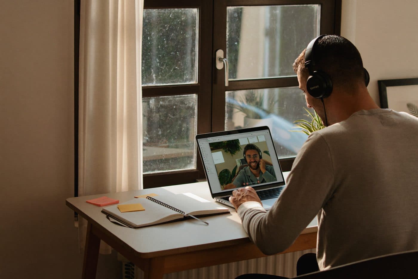 A man at a sunny desk with headphones on, mid video session, notebook open