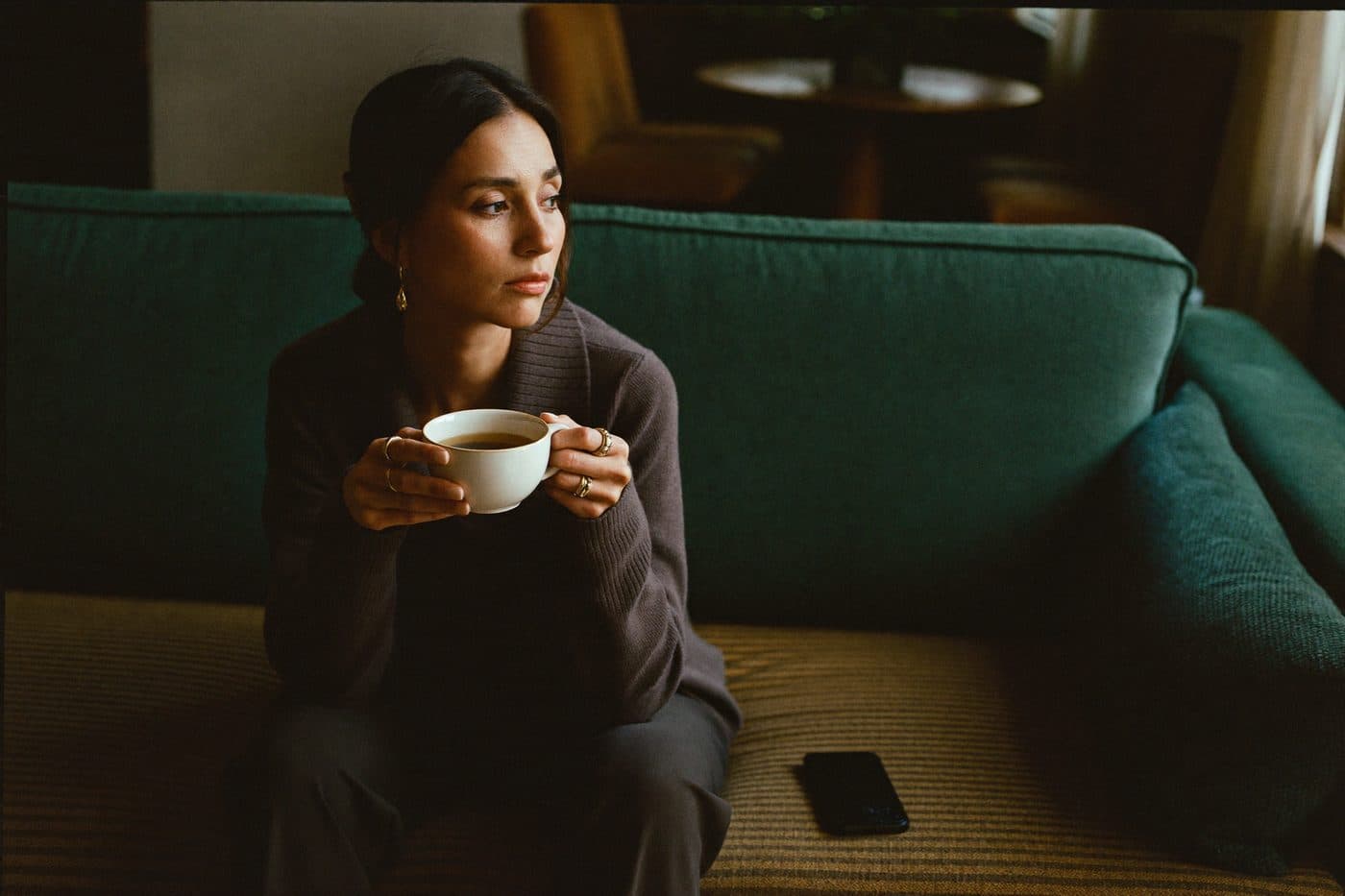 A woman alone on a green couch with a cup of tea, thoughtful, phone set aside