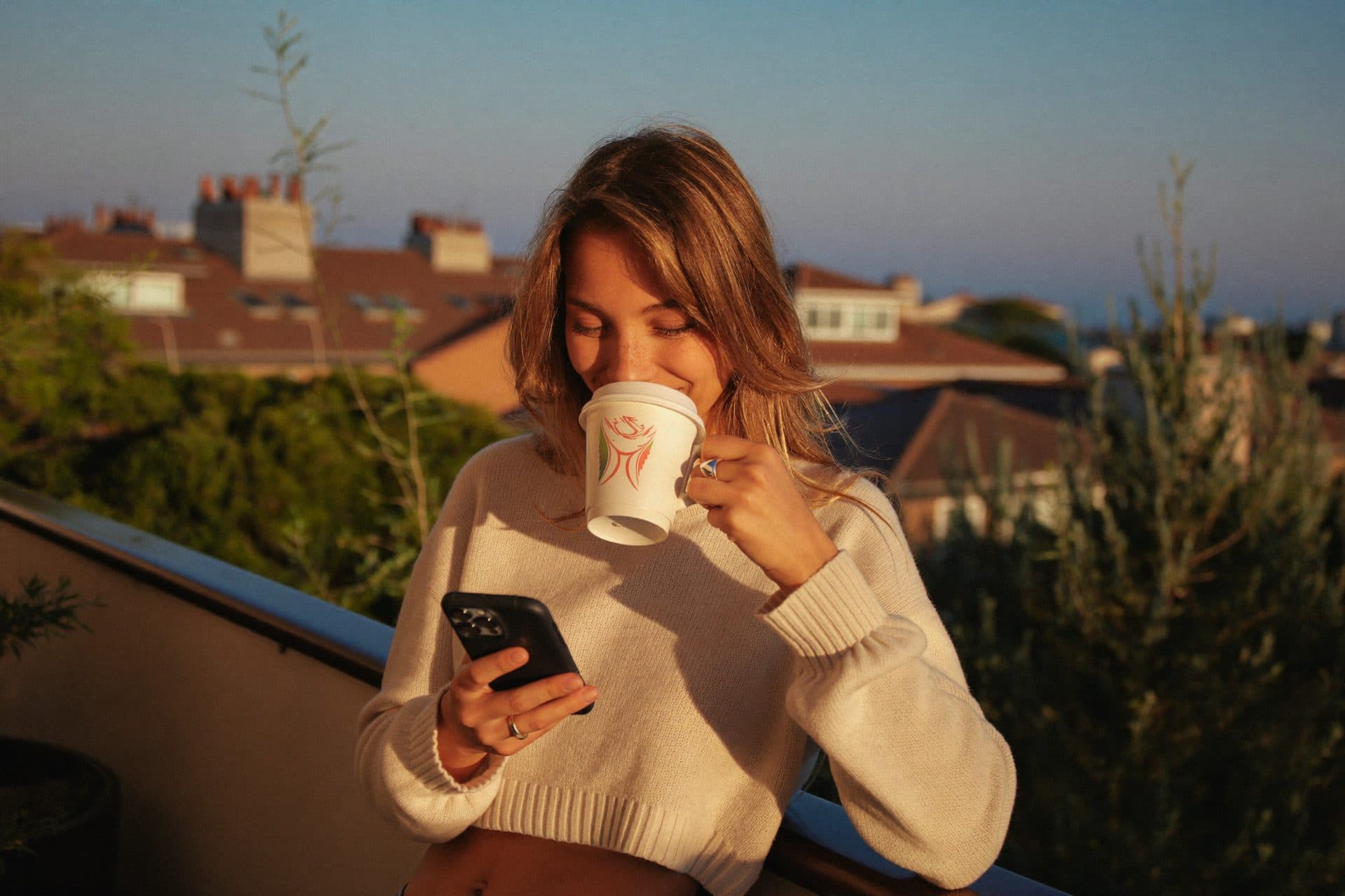 A woman on her balcony at golden hour, coffee in hand, glancing at her phone