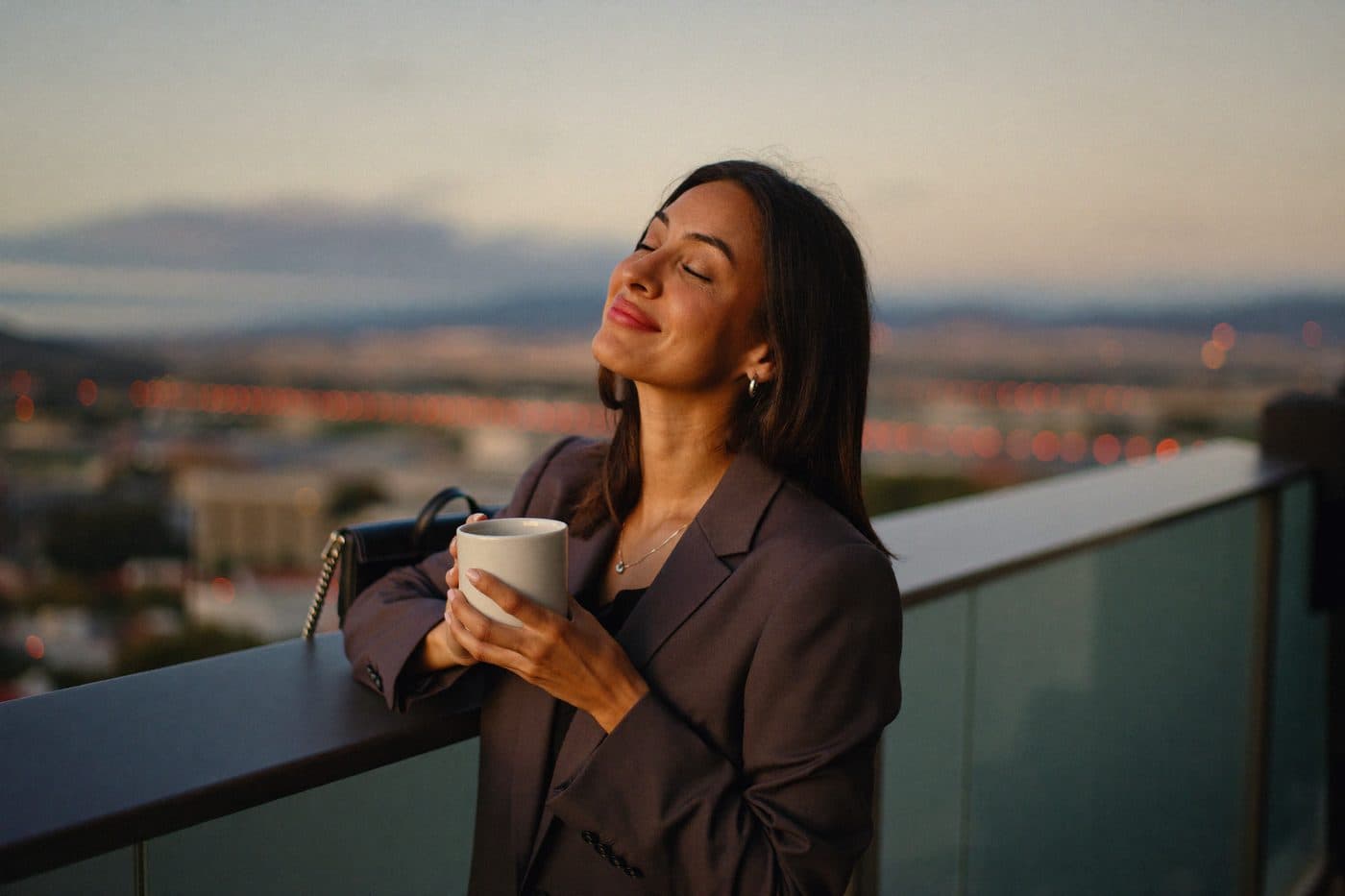 A professional on her balcony at dusk, mug in both hands, eyes closed, finally off the clock